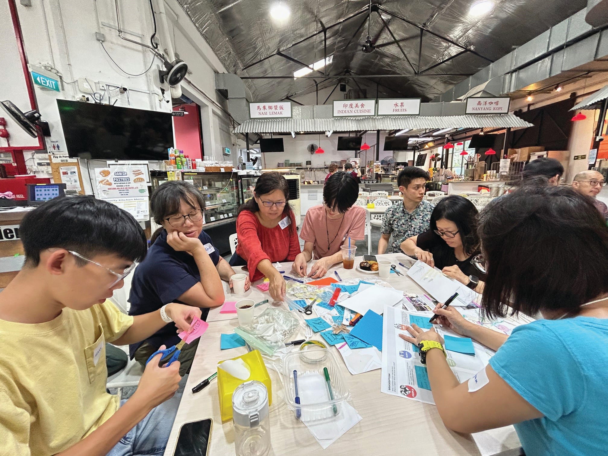 A group of people work on a craft project at a large table in a market-style food hall.